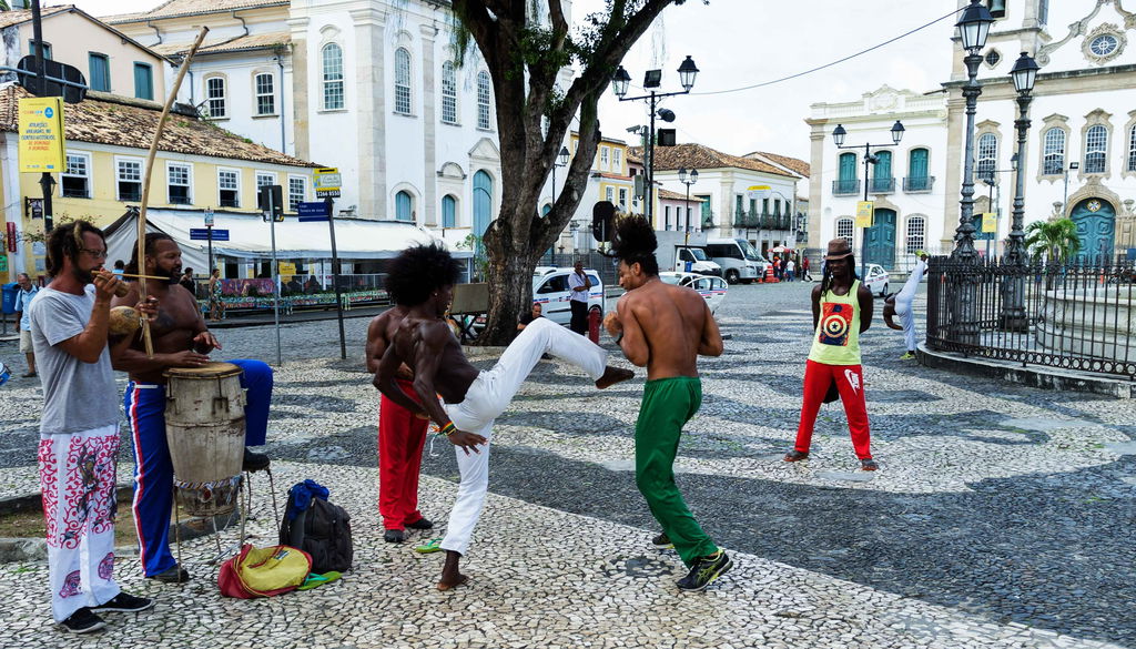 Na rua, dois homem sem camisa jogam capoeira, enquanto outros homens em roda tocam instrumentos.