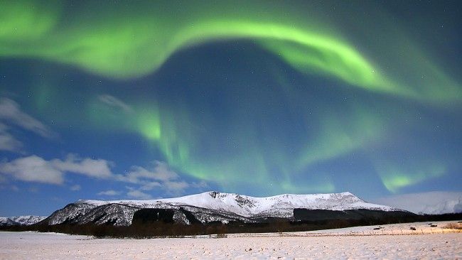 Paisagem de uma montanha coberta de neve e no céu azul acontece a aurora boreal em tom de verde.