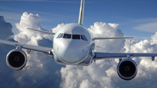 Avião branco, visto de frente, em céu azul sobrevoa nuvens brancas.
