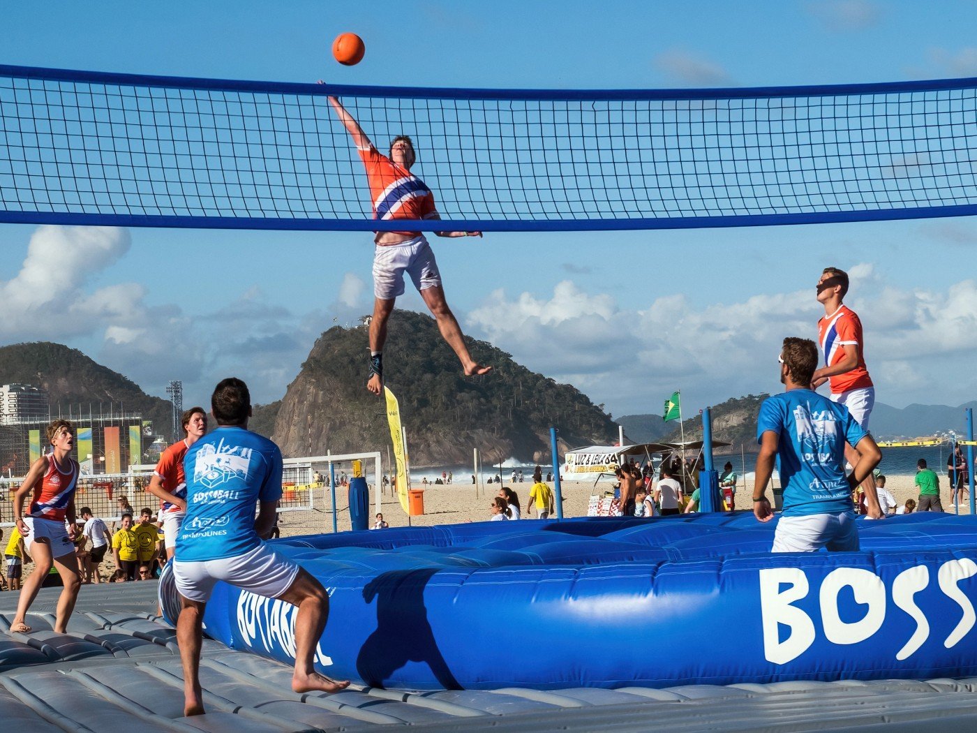 Jogadores sobre um trampolim inflável divido por uma rede, na praia, pulam e tentam jogar a bola na área do time adversário, que está preparado para defender.