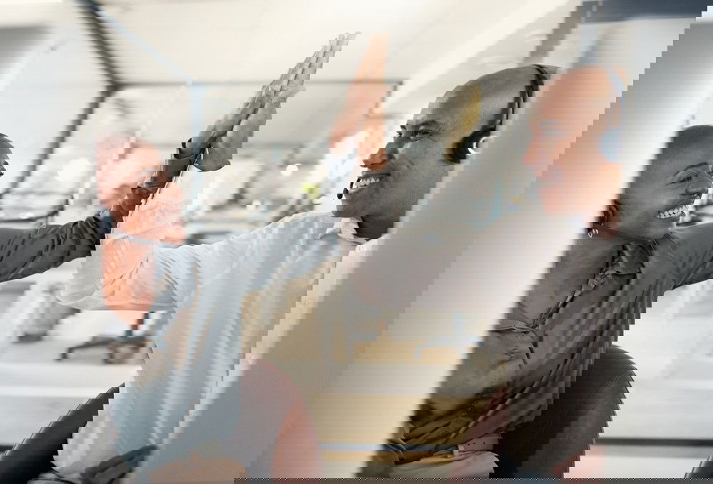 Homem e mulher negros vestidos em tons de branco e cinza, sentados um ao lado do outro, com fones de ouvido, fazem o cumprimento de “toca aqui” com as mãos.