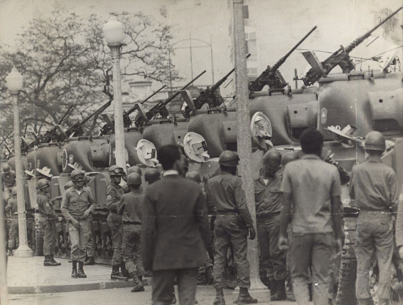 Tanques de guerra enfileirados a direita em uma rua, com as armas apontadas para cima, enquanto alguns homens caminham nas proximidades.