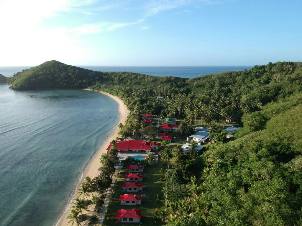 Praia de areias claras e água limpa, vista de cima, com floresta e montanha verde do lado direito.