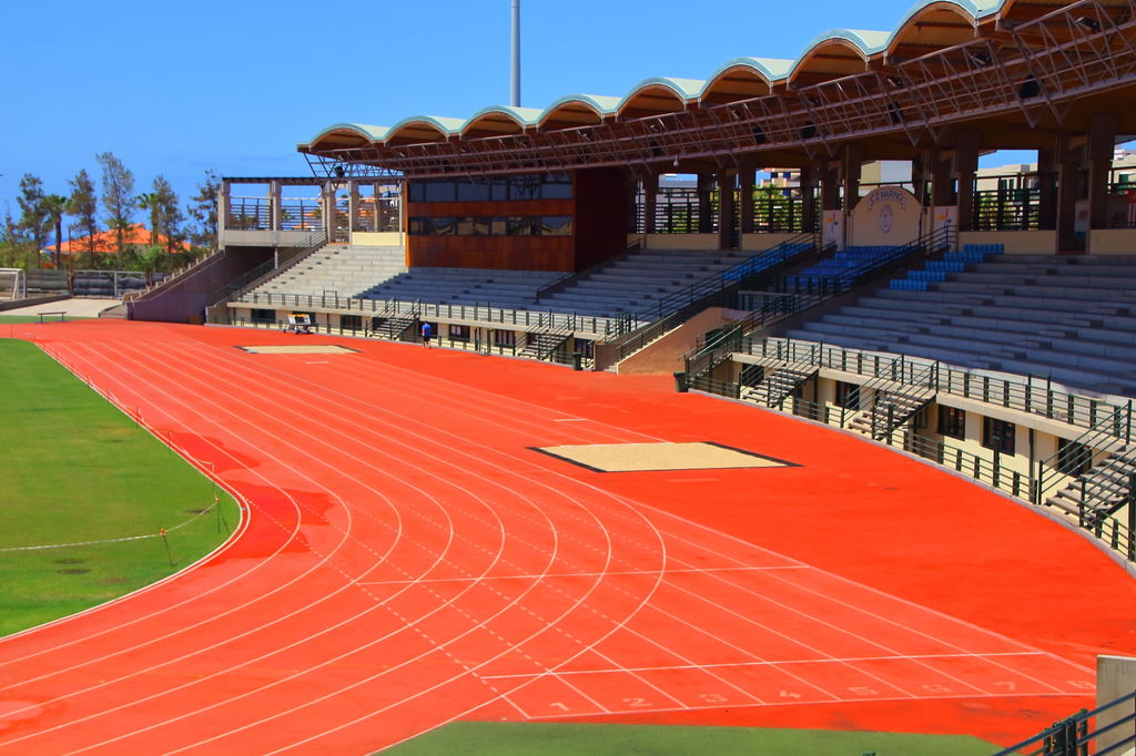 Estádio de atletismo, com destaque para a pista alaranjada de corridas e a arquibancada vazia em tons de azul.