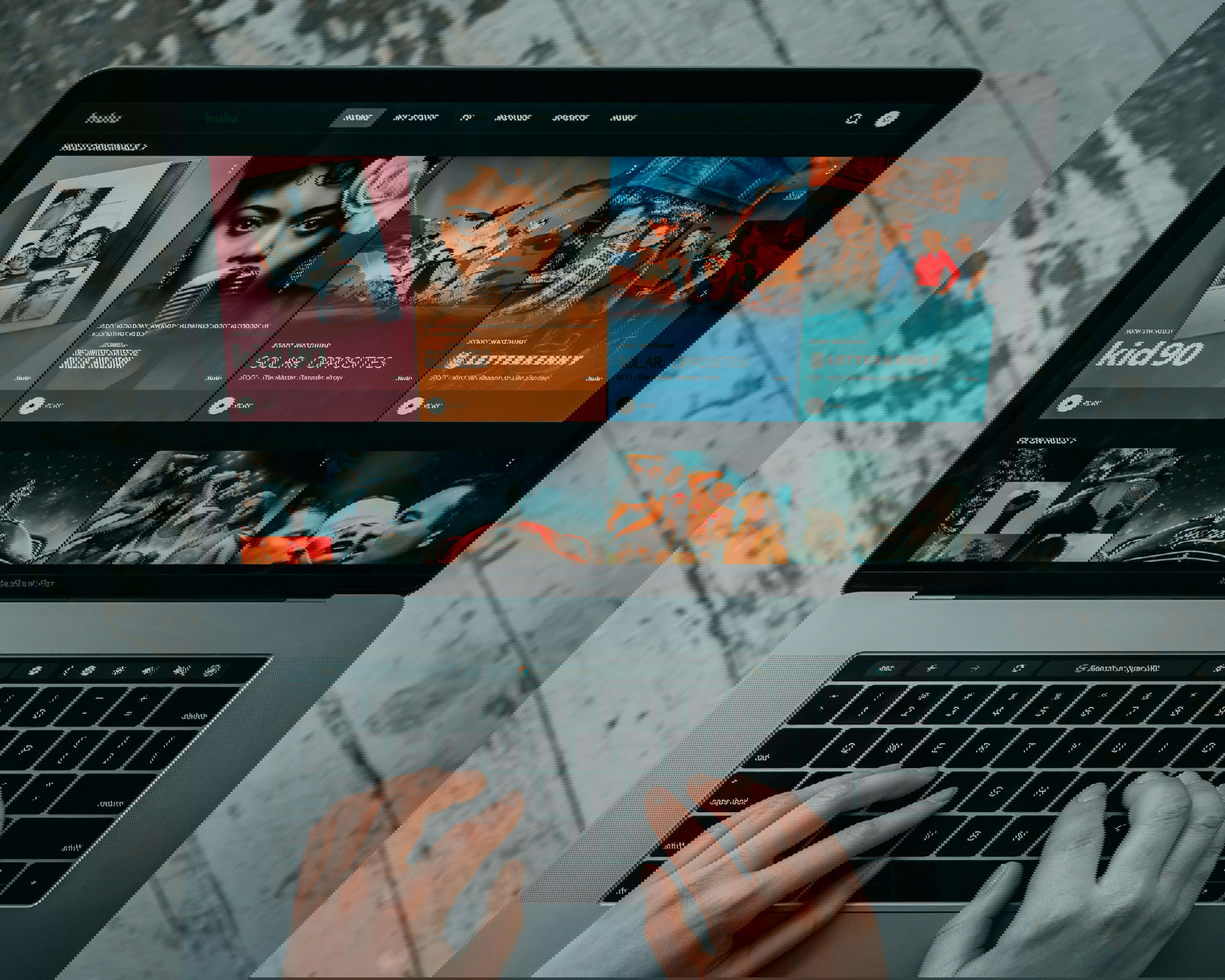 Mão branca digita sobre o teclado de um notebook em que na tela parece várias opções de filmes para streaming.