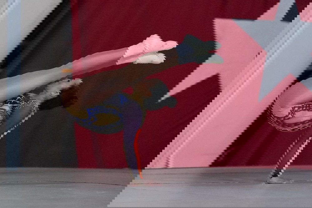 Ginasta, de maiô roxo, apoia-se com os braços esticados no chão enquanto inclina as pernas sobre o rosto.