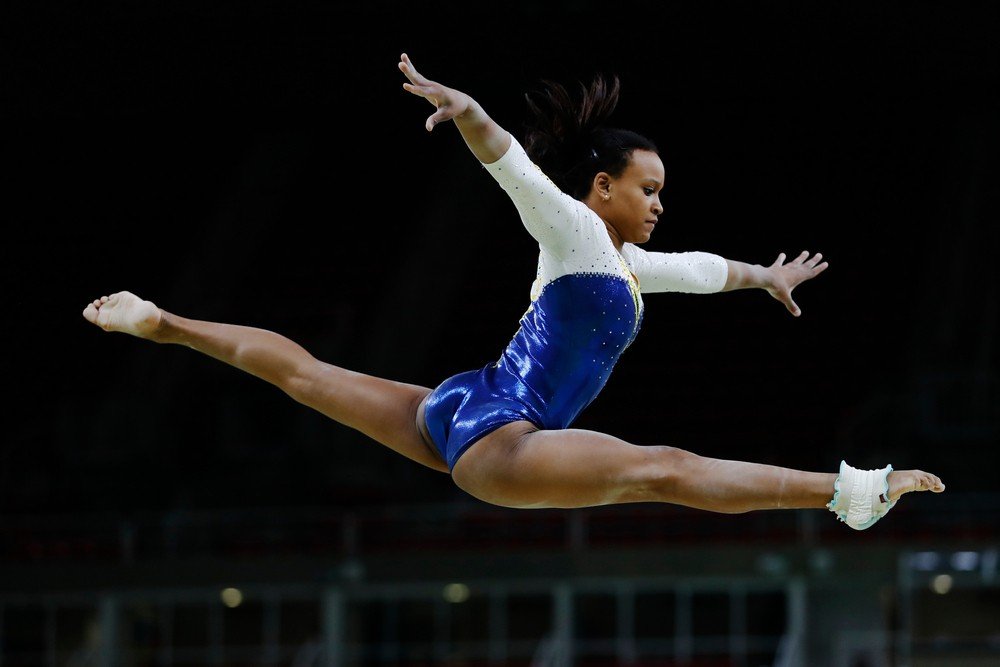 Atleta brasileira Rebeca Andrade, de maiô branco, faz salto com pernas e braços esticados no ar.