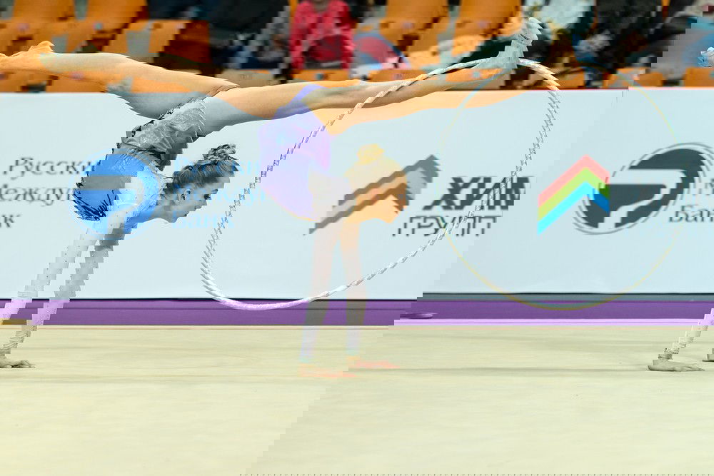 Ginasta rítmica se apoiando com os braços esticados no chão e pernas abertas para cima, segurando o arco com um dos pés.