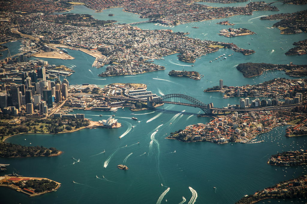 Porto de Sydney na Austrália, visto de cima, mar azul e barcos se movimentando abaixo de grandes pontes.