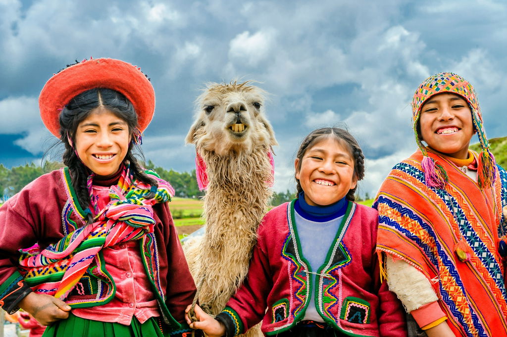 Duas meninas, um menino e uma lhama, com roupas tradicionais peruanas sorriem.