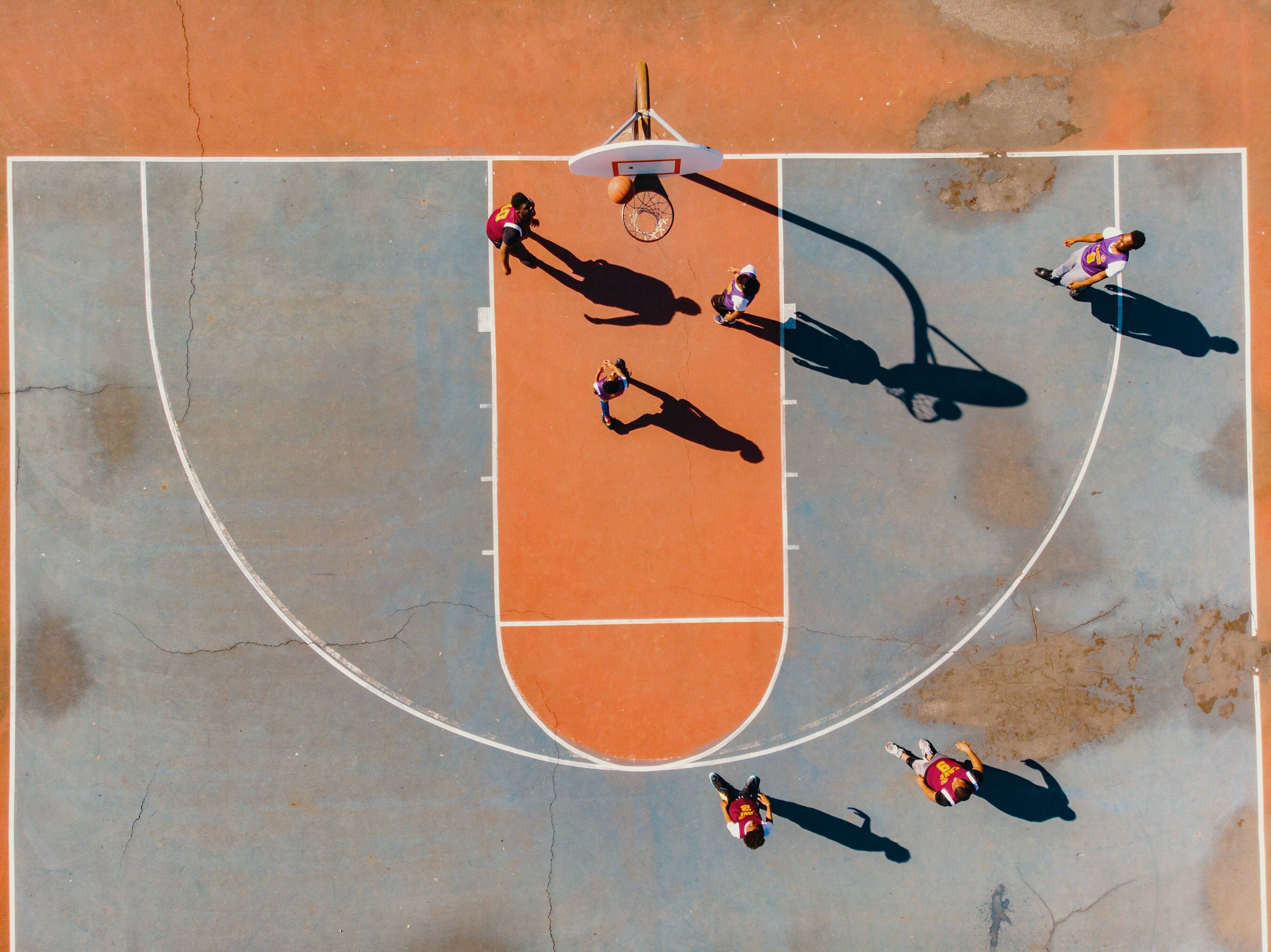 Vista superior de uma quadra de basquete, na qual seis pessoas jogam e uma marca cesta.
