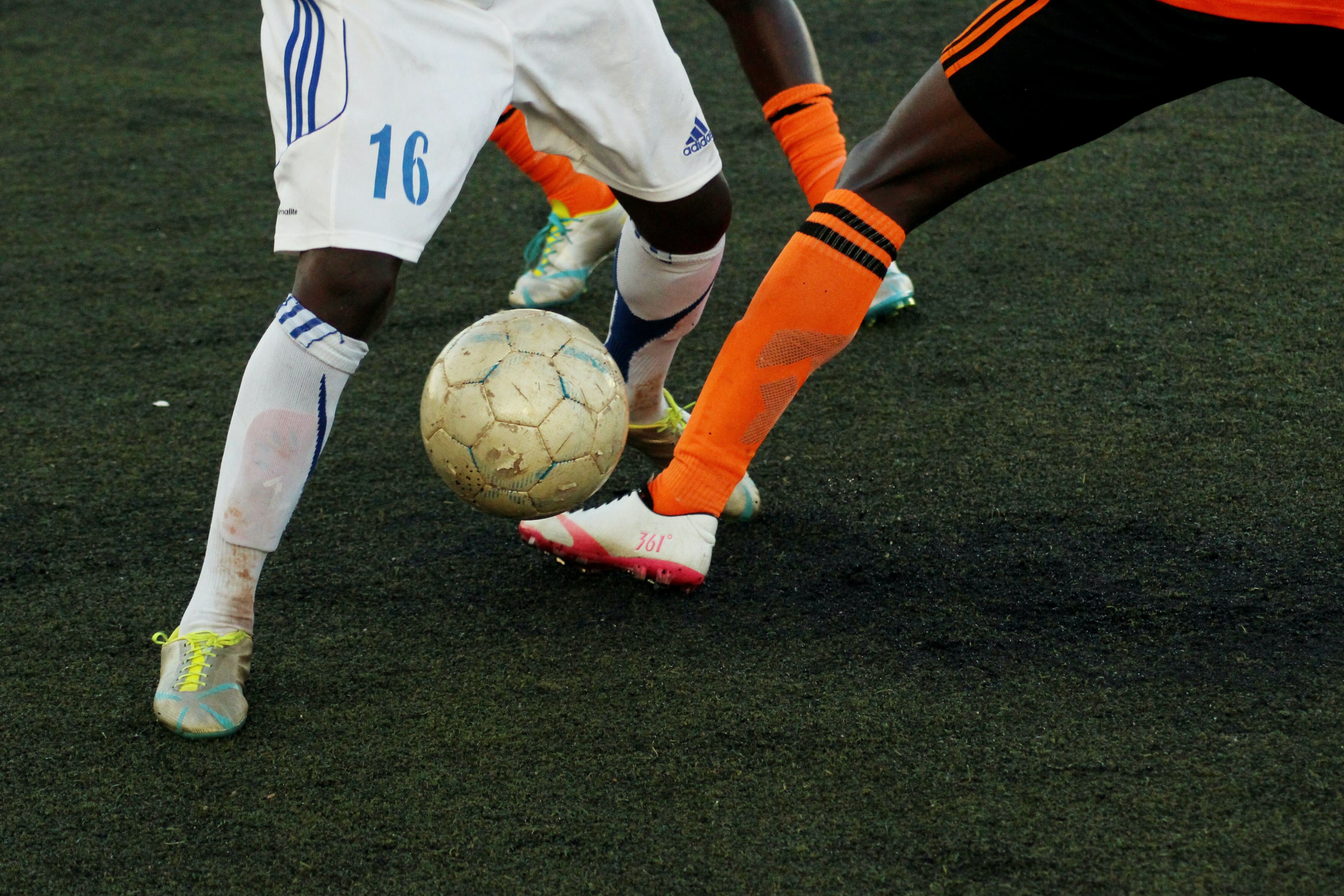 Em gramado do campo de futebol, ângulo mostra as pernas de dois jogadores, enquanto um tenta driblar o outro.