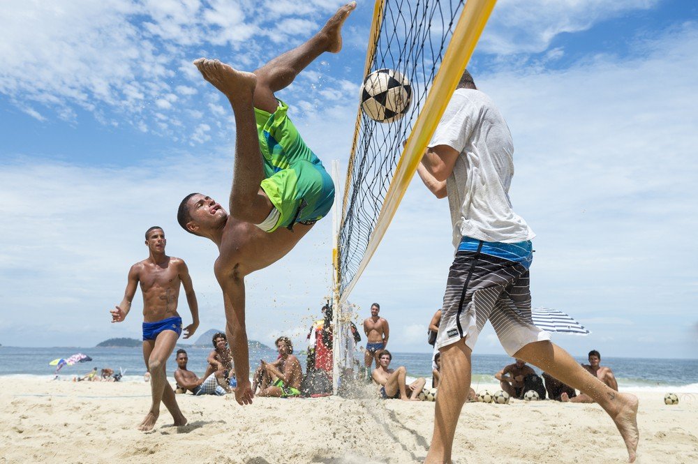 Três homens jogam futevôlei na praia, em um dia ensolarado, enquanto um grupo de pessoas assiste.