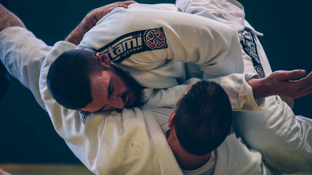 Dois atletas em trajes brancos estão juntos no chão, em uma manobra de jiu jitsu brasileiro.