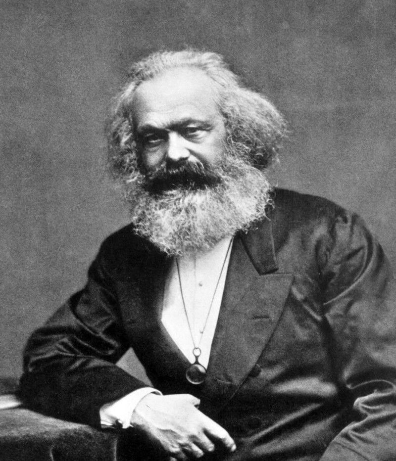 Filósofo Karl Marx, em preto e branco, homem branco de cabelo branco ligeiramente comprido, assim como sua barba, usa um terno preto, está sentado e olha para a câmera sem sorrir.