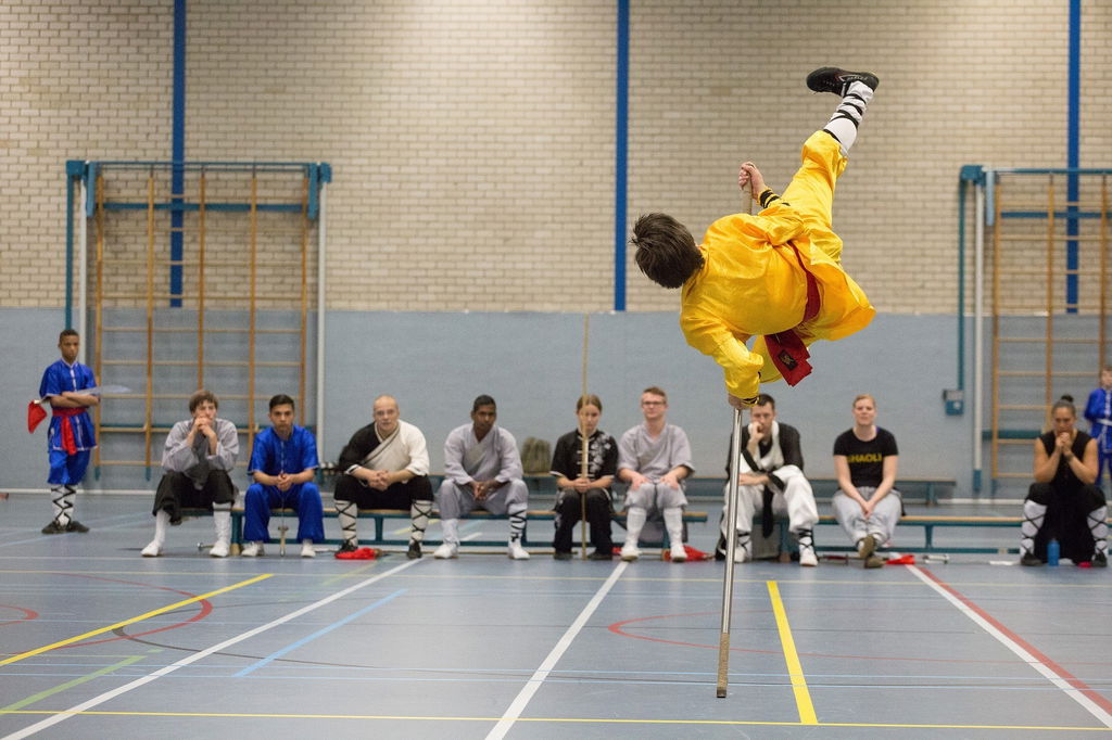 Homem em roupas amarelas está no ar, apoiado por uma vara cinza, em um golpe de kung fu.