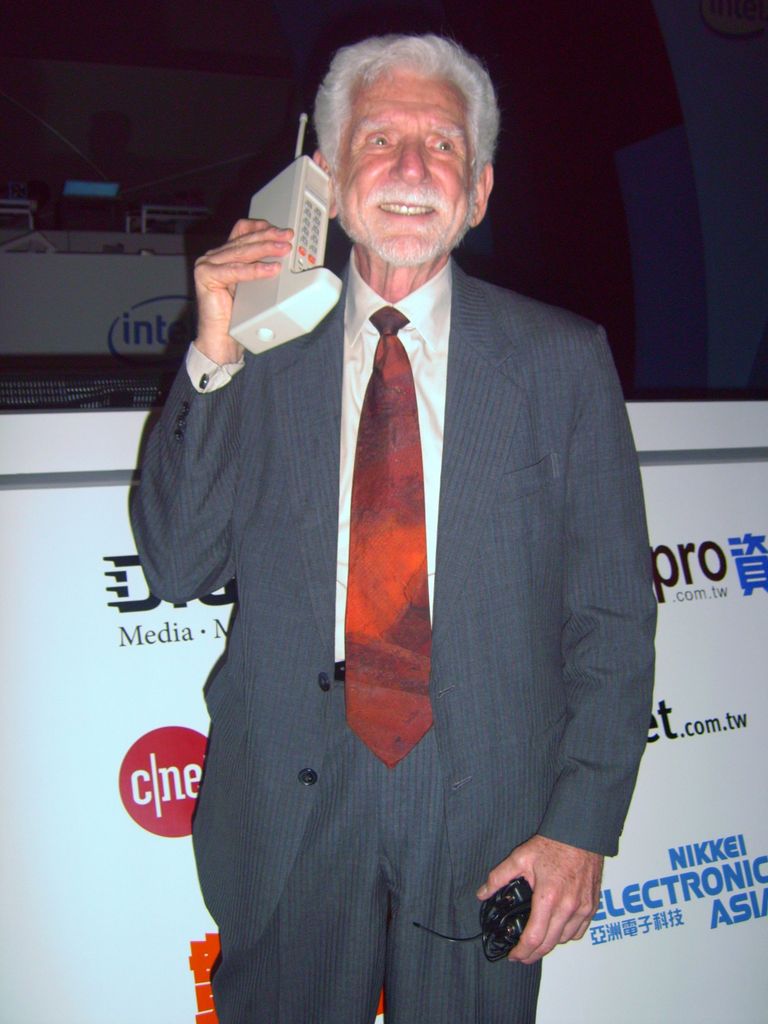 Senhor de cabelos brancos vestido de terno cinza sorri enquanto segura perto do rosto um celular antigo.