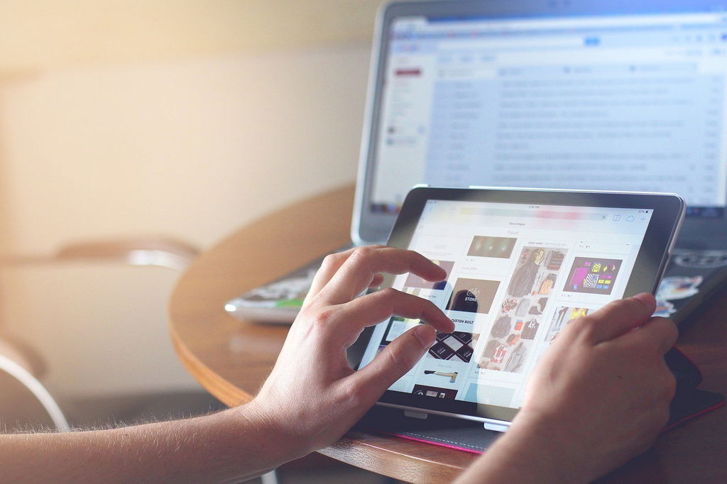Mãos femininas brancas usam um tablet em primeiro plano, atrás um computador ligado.