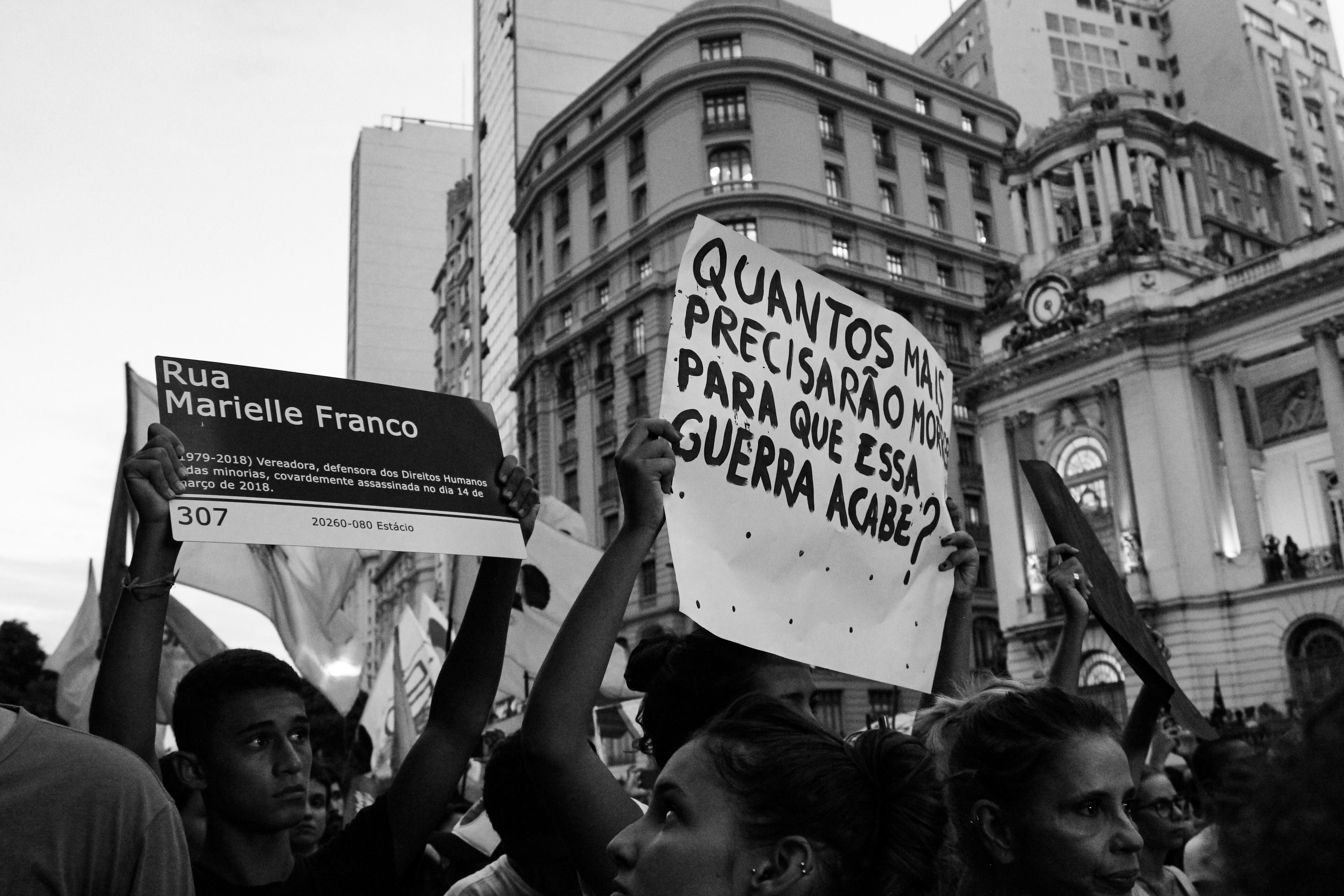 Protesto, em preto e branco, em que manifestantes levantam placas sobre violência contra a população negra.