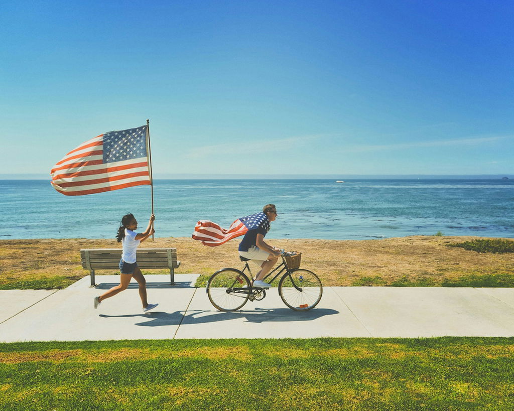 Homem anda de bicicleta com a bandeira dos Estados Unidos presa às costas, e menina corre atrás da bicicleta segurando outra bandeira americana.