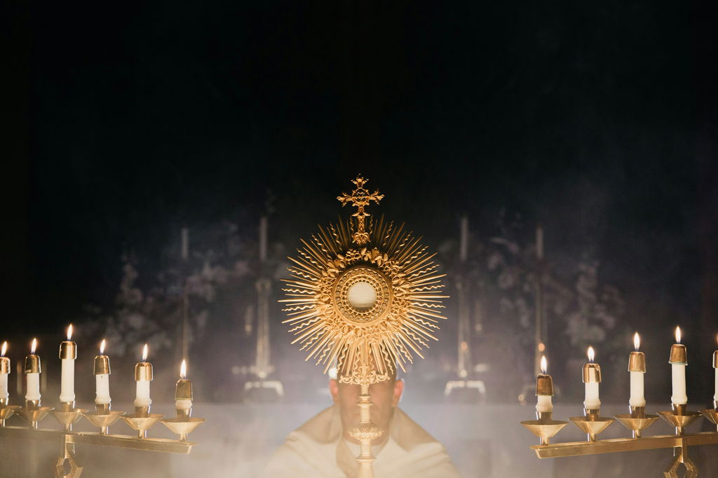 A hóstia consagrada vista dentro de uma redoma dourada, com padre e igreja ao fundo.