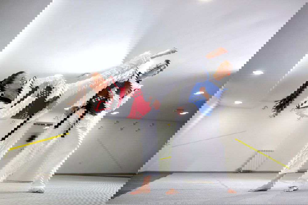 Homem e mulher de quimonos brancos e proteções de tronco azul e vermelha lutam  taekwondo.