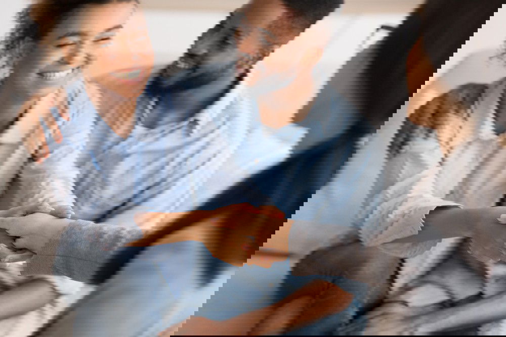 Homem e mulher, vestidos de tons claros, sentados um ao lado do outro, cumprimentam outra mulher, sentada a frente.